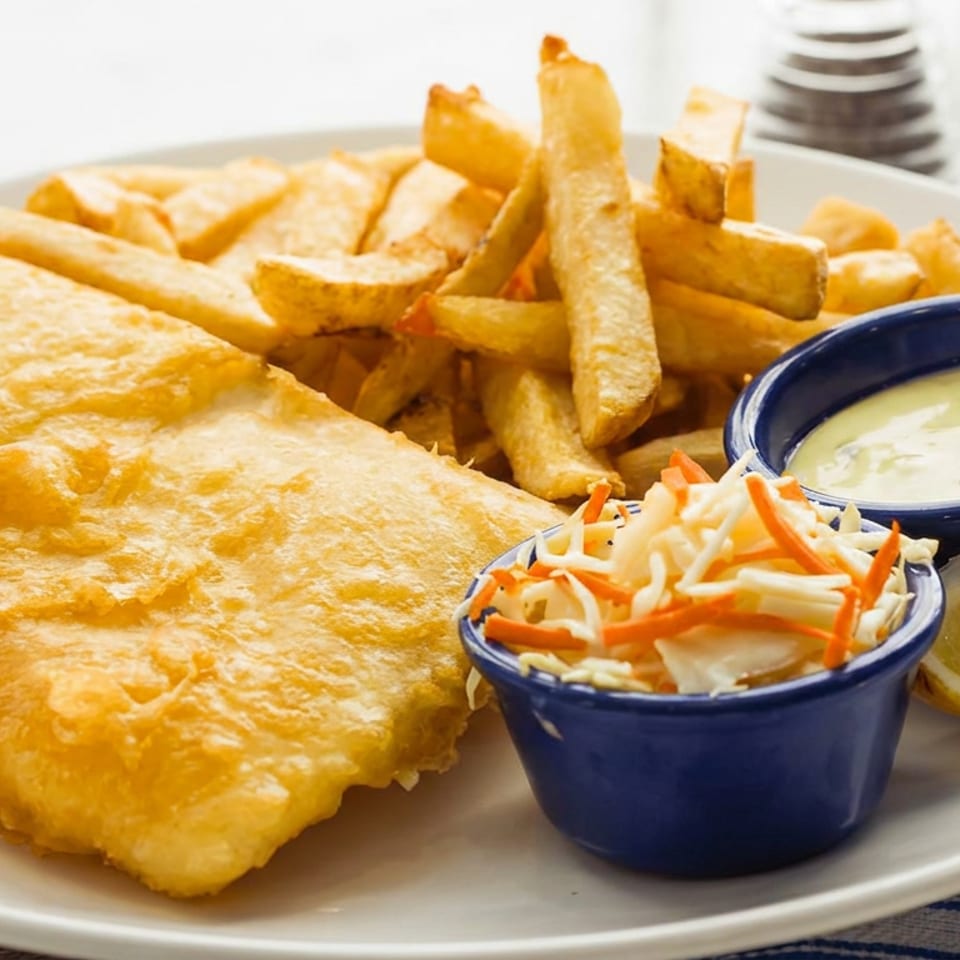 Golden battered fish with French fries, coleslaw, tartar sauce, and lemon wedge on a white plate.